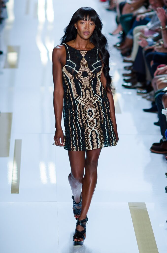 Naomi Campbell walking a fashion runway in an intricate black and gold dress
