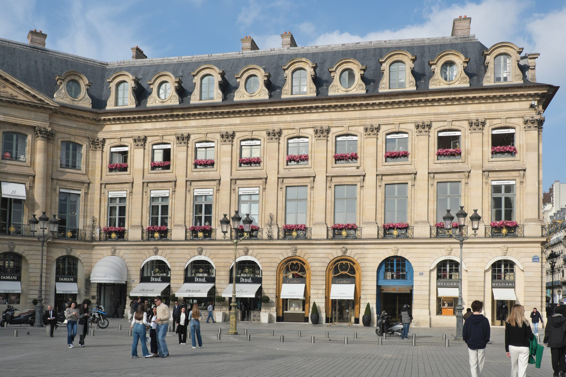 Pedestrians beneath pink flower boxes on sunny Place Vendôme near Rue Saint-Honoré