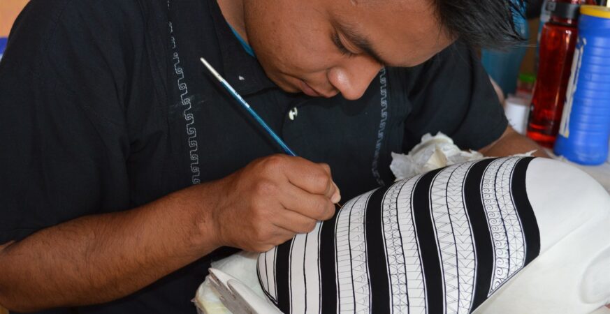 Un jeune artisan peint les détails d’un alebrije à San Martín Tilcajete : visage, main et créature replacent l’auteur humain au centre de l’image.