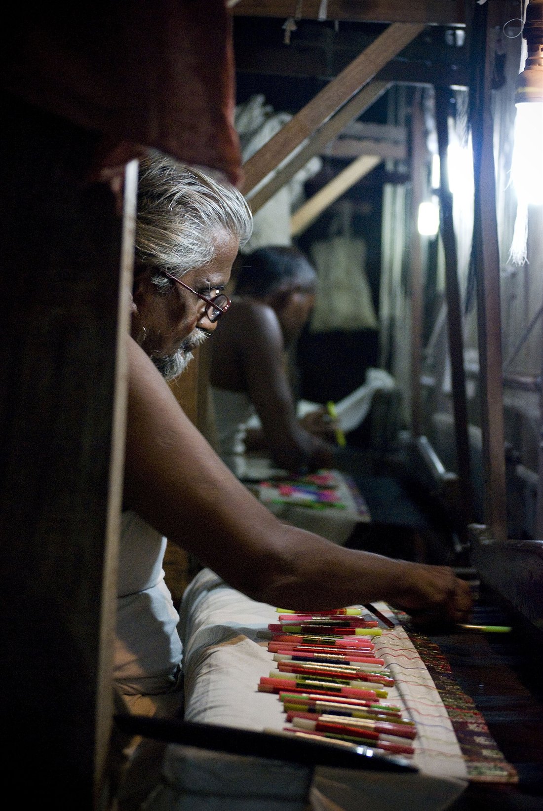 A Varanasi silk weaver works beside coloured weft bobbins—the slow labour hidden by the pattern’s finished precision.