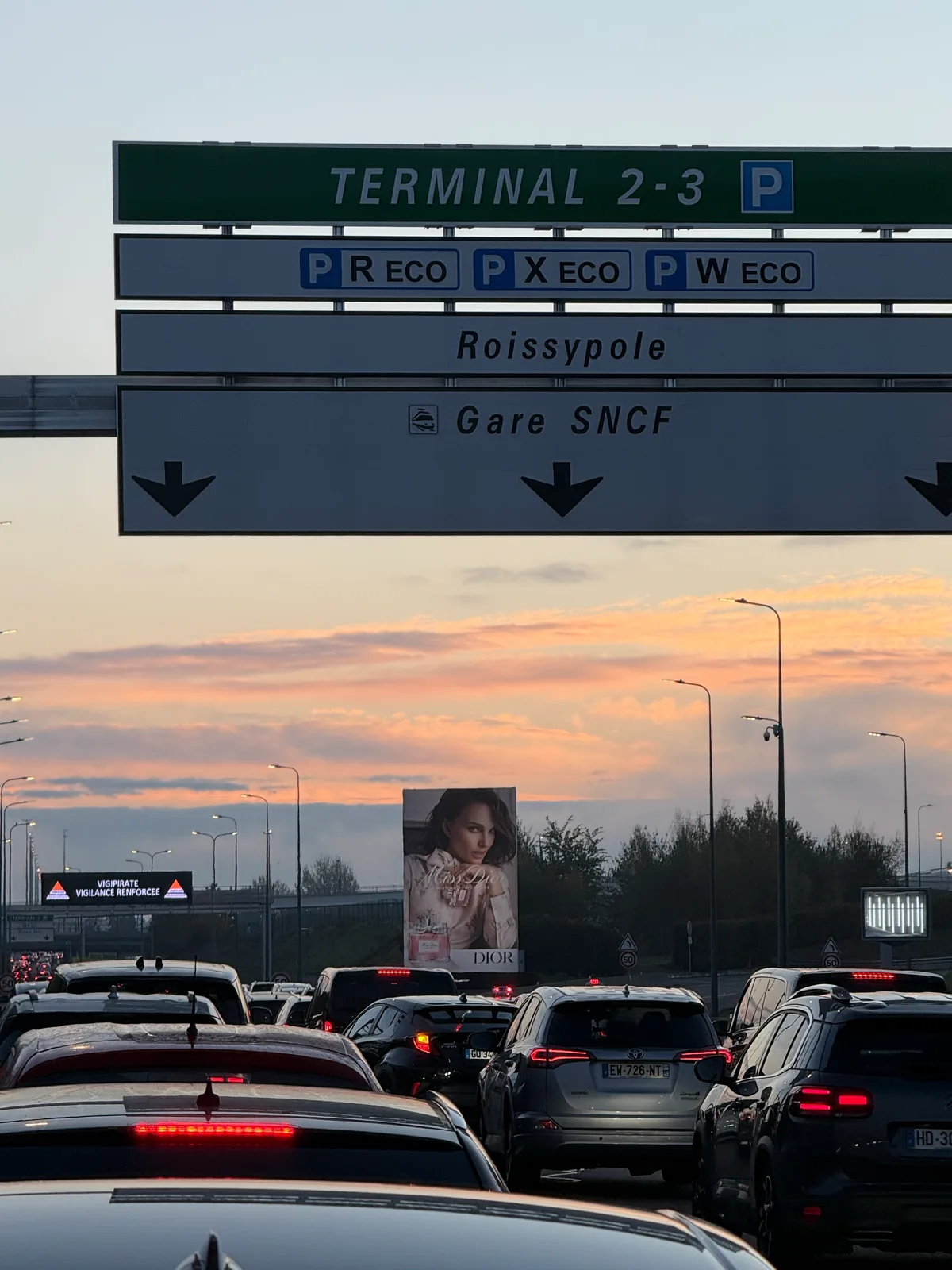 ازدحام صباحي عند مداخل مجمّع Terminal 2 في مطار شارل ديغول، مع لوحات توجيه إلى Roissypôle ومحطة SNCF.