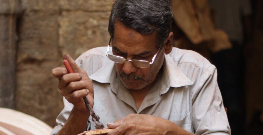 Un facteur de oud de la rue Mohamed Ali au Caire façonne la caisse à côtes : visage, main et matière replacent l’artisan au centre du récit.