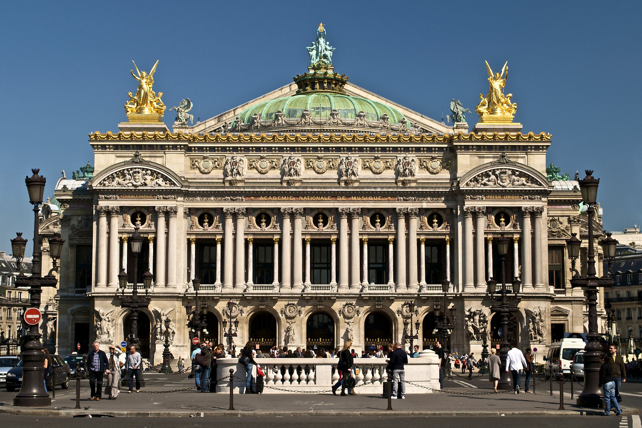 Bright frontal view of the Palais Garnier façade in Paris with incidental pedestrians on Place de l’Opéra